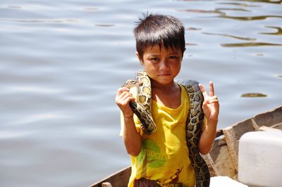 Portrait of cute boy holding snake while showing peace sign on boat against lake