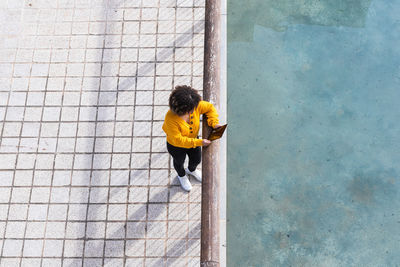 From above remote view of female with afro hairstyle standing near metal railing in urban area of city while using a tablet device