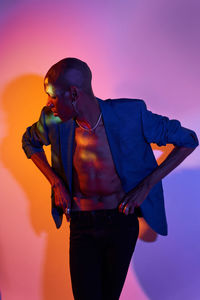 Confident bald african american slim male model with necklace in trendy outfit standing at wall with glowing lights in modern studio