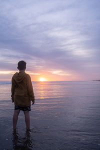 Rear view of man standing at beach against sky during sunset