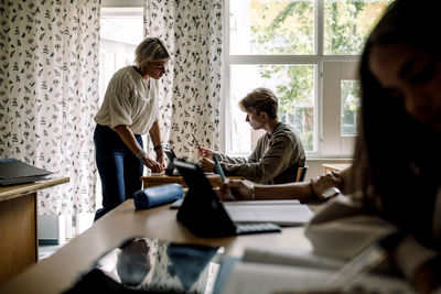 Female teacher helping teenage boy while studying in classroom