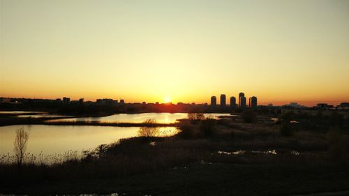 Silhouette buildings by river against sky during sunset