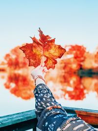 Cropped image of person holding orange maple leaf at lake against clear sky