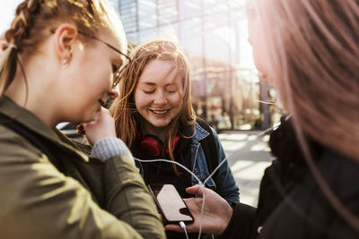 Teenage girl looking at friends listening music through mobile phone in city