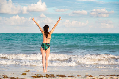 Rear view of woman wearing bikini while standing with arms raised at beach