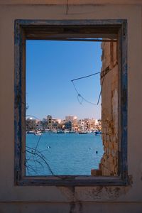 Buildings by sea against clear blue sky seen through window