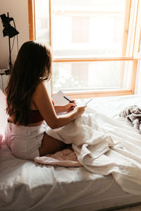 Woman writing in book while sitting on bed at home