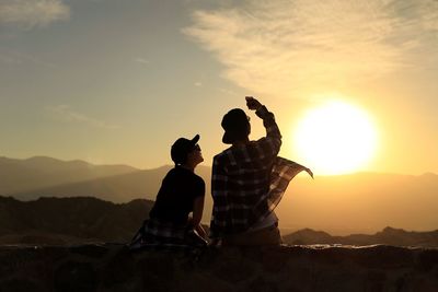 Rear view of couple taking selfie while sitting on retaining wall against sky during sunset