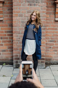 Cropped hands of young man photographing fashionable woman standing against brick wall