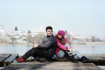 Smiling couple sitting on pier over lake against sky