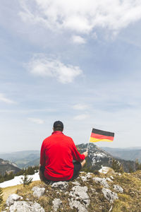 Rear view of man with flag sitting on rock against sky