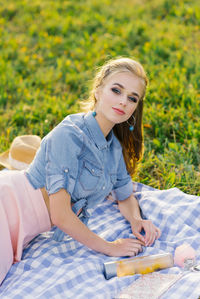 Stylish young woman with bright makeup in a denim shirt is resting on a plaid in the open air