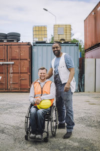 Portrait of smiling construction worker standing by colleague sitting in wheelchair at site