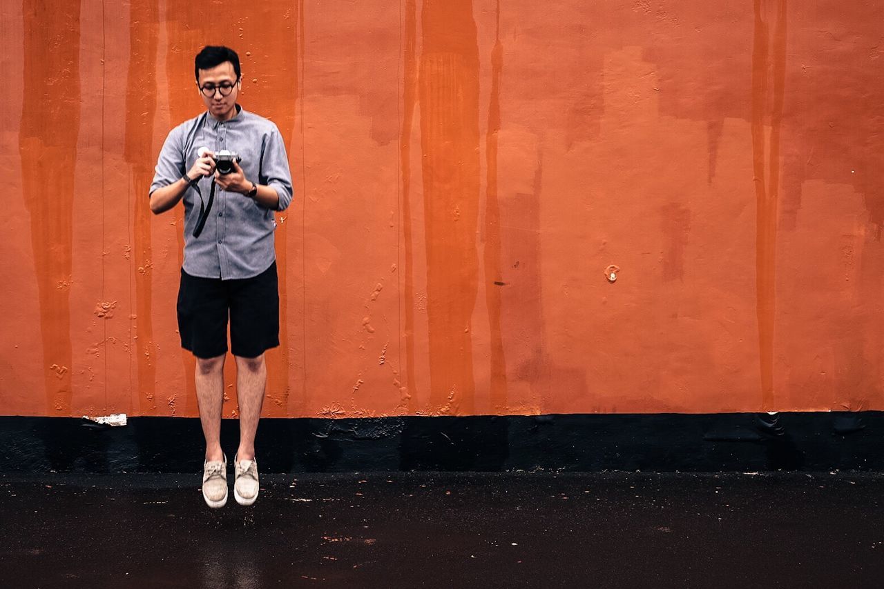 FULL LENGTH OF MAN HOLDING MOBILE PHONE STANDING AGAINST WALL