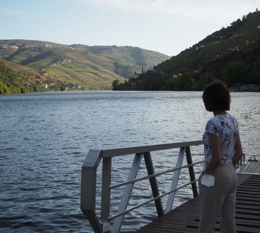 REAR VIEW OF MAN STANDING ON RAILING BY LAKE AGAINST MOUNTAINS