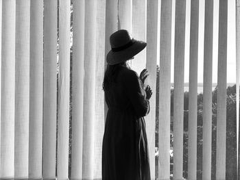 Black and white photo of woman wearing hat standing near the blinds