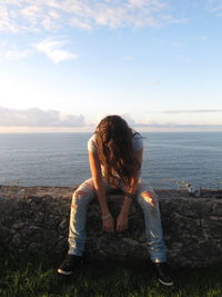 Woman covering face with hair while sitting on retaining wall by sea