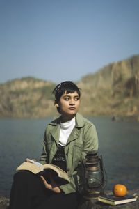 Young woman looking away while holding book sitting by lake