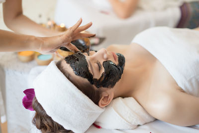 Beautician applying coal facial mask on face of young woman in spa