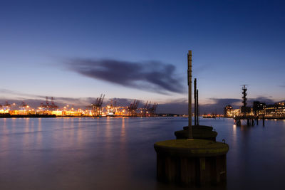 Wooden posts in sea by illuminated city against sky at dusk