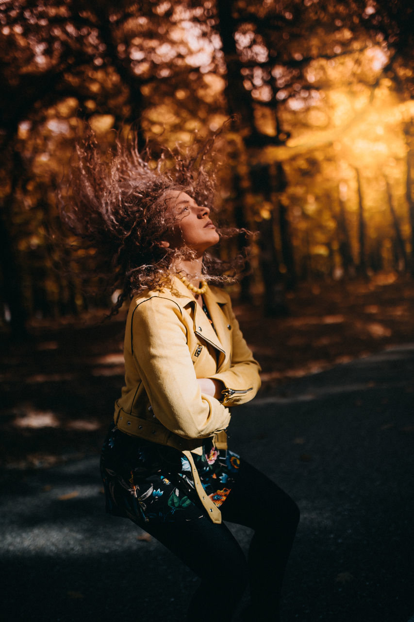 one person, tree, adult, women, nature, autumn, sunlight, forest, hairstyle, lifestyles, young adult, female, leisure activity, plant, clothing, person, light, child, emotion, smiling, darkness, land, outdoors, spring, side view, casual clothing, childhood, happiness, curly hair, looking, sunset, beauty in nature, portrait, relaxation, road, long hair, motion, fashion, full length, city, arts culture and entertainment