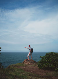Man pointing while standing on rock by sea against cloudy sky