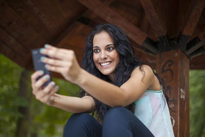 Low angle view of cheerful young woman using phone while sitting in gazebo