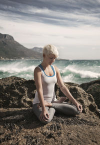 Blond woman meditating while practicing lotus position yoga on rock formation at beach