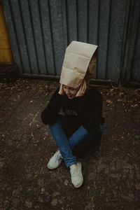 High angle view of woman wearing paper bag sitting against wall