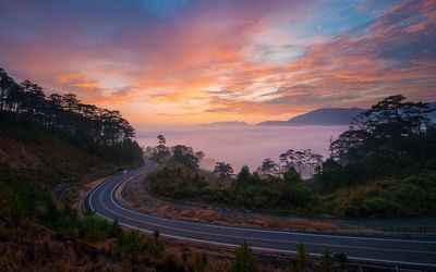 Road by trees against sky during sunset