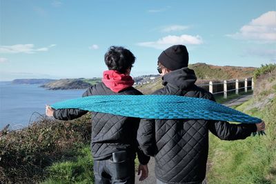 Rear view of friends holding scarf while standing on hill by sea against sky