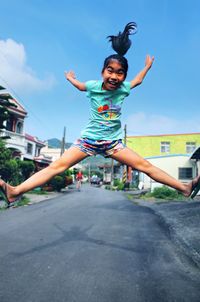 Portrait of happy girl with arms outstretched jumping on road against blue sky