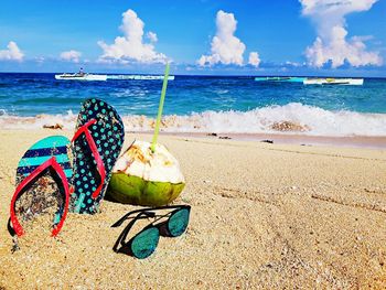 Slippers with sunglasses and coconut on sand at beach during sunny day