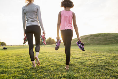 Rear view of mother and daughter holding sports shoes while walking on grass at park during sunset