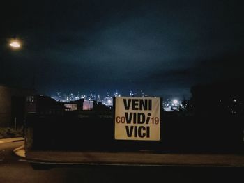 Information sign on road by buildings against sky at night