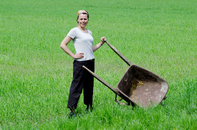 Portrait of confident woman with hand on hip and wheelbarrow standing on farm