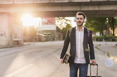 Young man on the move with skateboard, rolling suitcase and headphones