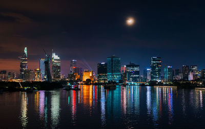 Illuminated buildings by river against sky at night