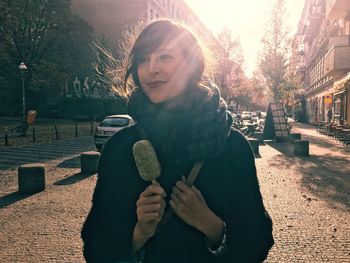 Smiling young woman holding ice cream against buildings on sunny day