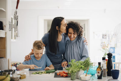 Smiling woman kissing partner while son cutting cucumber in kitchen