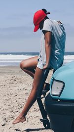 Side view of woman sitting on car at beach against sky