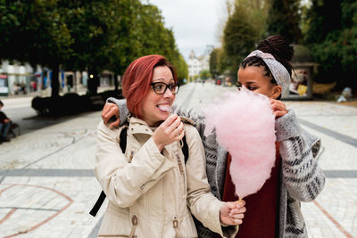 Stylish cheerful multiethnic girl friends walking embracing and eating pink candy cotton in street