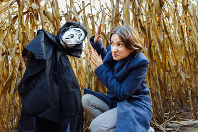 Woman close up with hands next to creepy scarecrow with mask of death in cornfield on halloween