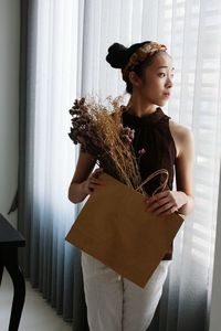 Young woman holding paper bag with bouquet while standing by window at home