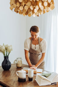 Housewife preparing dough standing at domestic kitchen, wearing apron