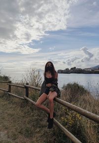 Full length of young woman wearing mask sitting on railing against sky