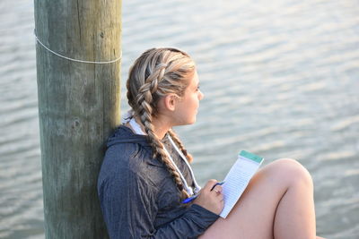 High angle view of thoughtful teenage girl writing on note pad while sitting by lake