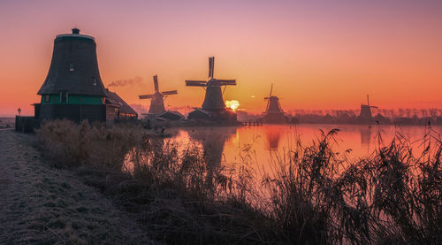 Traditional windmill by lake against sky during sunset