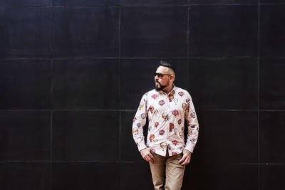 Bearded man wearing sunglasses leaning on black wall, looking aside