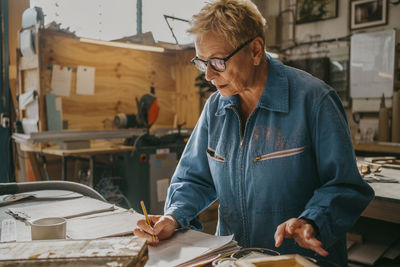 Senior female carpenter wearing eyeglasses preparing document at workshop